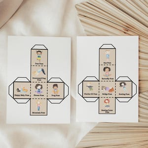 Op de afbeelding: Twee witte kaarten met illustraties van yogahoudingen. Elke kaart heeft een kubusontwerp met tekeningen van kinderen die houdingen demonstreren zoals Boomhouding, Neerwaartse Hondhouding en Gelukkige Babyhouding. De kaarten liggen op een beige oppervlak.