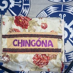 May include: A floral fabric with red and cream-colored flowers and green leaves. The fabric has a tan banner with the word "CHINGÓNA" in sparkly purple letters. The background is a blue and white patterned fabric.