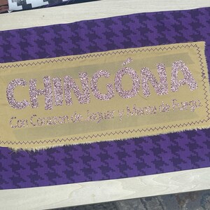 May include: A rectangular patch with the word "CHINGÓNA" in glittery pink letters on a tan background. The patch is sewn onto a purple houndstooth fabric. The text "Con Corazon de Jaguar y Mente de Fuego" is below.