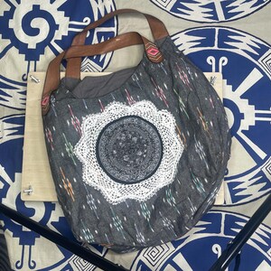 May include: A gray tote bag with a white lace applique and brown leather straps. The bag features a patterned design with colorful accents. The background includes a patterned fabric with blue and white circular designs.