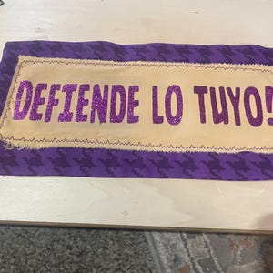 May include: A rectangular fabric piece with the Spanish text "DEFIENDE LO TUYO!" in glittery purple letters. The text is on a tan background, framed by a purple houndstooth pattern. The edges are stitched with a decorative thread.