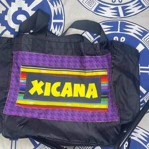 May include: Black tote bag with a purple houndstooth pattern and a rectangular patch featuring the word "XICANA" in yellow. The patch has a colourful striped border. The bag has black handles.