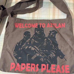 May include: Brown canvas tote bag with a shoulder strap. The bag features the text "WELCOME TO AZTLAN" and "PAPERS PLEASE" in red. A black graphic of three figures in military gear is printed on the front.
