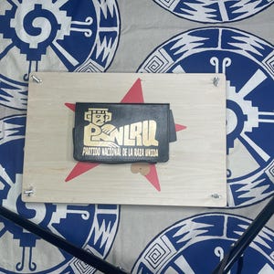 May include: A black wallet with gold lettering and a graphic design is displayed on a wooden board with a red star. The board is secured with silver hardware. The background features a patterned fabric with blue and white circular designs.