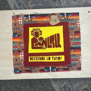 May include: A wooden frame holds a fabric piece with a central yellow rectangle featuring a brown figure and the letters "NRLU." The text "DEFIENDE LO TUYO!" is below. The fabric has a brown and red pattern.