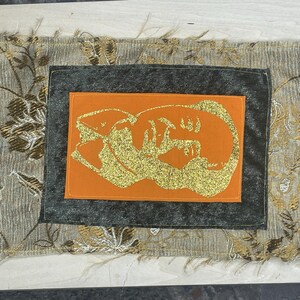 May include: A rectangular textile piece with a layered design. The outer layer is a gray fabric with a floral pattern in gold and brown. Inside is a dark gray square, and an orange square with a gold glitter design.