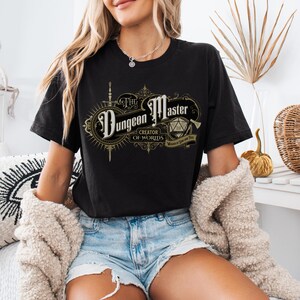 Puede incluir: Camiseta negra con un diseño gráfico dorado que dice "The Dungeon Master Creator of Worlds Master of Dice and Time".