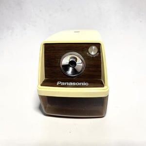 May include: A vintage Panasonic electric pencil sharpener. The sharpener has a cream-colored top and base with a dark wood-grain front panel. The word "Panasonic" is printed on the front. The sharpener has a silver-colored dial and an "AUTO-STOP" button.