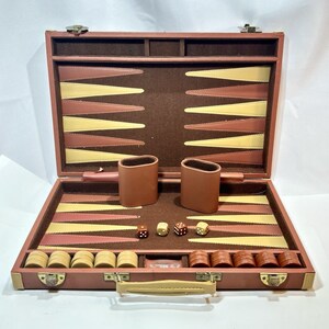 May include: A brown backgammon set in a briefcase-style case with a brown felt interior. The board features alternating light cream and brown triangular points. Included are two brown leather dice cups, four dice, and a set of cream and brown checkers.