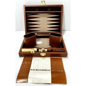 May include: Open brown leather backgammon set with a beige and brown playing surface. Includes dice, ivory-colored game pieces, and two brown leather dice cups. The set is displayed with an instruction booklet that reads "BACKGAMMON".
