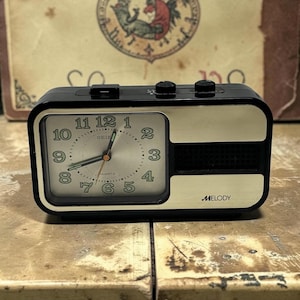 May include: A rectangular black and cream Seiko quartz alarm clock with a silver face, green Arabic numerals, and three hands. The clock features a speaker grille on the right side, the word MELODY printed in black, and three black control buttons on top.
