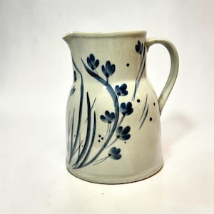 May include: A ceramic pitcher with a light cream-colored glaze featuring hand-painted blue floral motifs and slender green leaves. The vessel has a wide base, a curved handle on the right side, and a distinct pouring spout at the top rim.