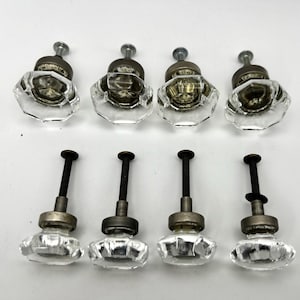 May include: A set of eight clear glass cabinet knobs with an octagonal faceted shape. Each knob features a metallic base with a dark finish and a threaded metal bolt extending from the back for installation, arranged in two rows of four pieces.