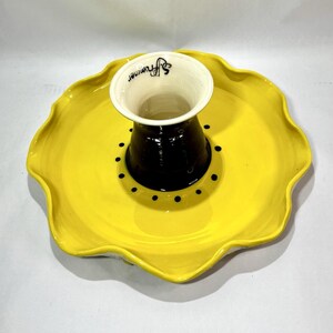 May include: A bright yellow ceramic serving dish with a scalloped edge and a central dark brown cylindrical pedestal. The pedestal features a white interior rim with the black text S. Frommer printed on it, surrounded by a ring of black dots.