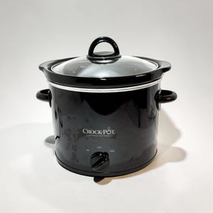 May include: A black Crock-Pot slow cooker with a clear glass lid and a central handle. The front features the white Crock-Pot logo, with a black control knob below labeled with Off, Low, and High settings. It has two side handles for lifting.