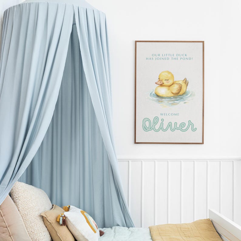 May include: A framed nursery print features a watercolor illustration of a yellow duckling in water. The text reads "OUR LITTLE DUCK HAS JOINED THE POND!" and "WELCOME Oliver" in a whimsical font. The print is displayed above a crib with a blue canopy.