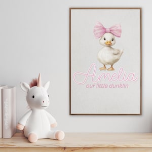 May include: A framed print featuring a watercolor illustration of a white duckling with a pink bow. The name "Amelia" is written in pink script, with "our little dunklin" below. A white unicorn plush toy sits on a wooden surface.