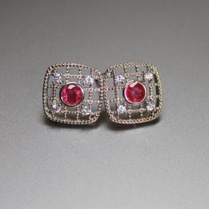 May include: A pair of square stud earrings featuring a silver-toned metal grid frame. Each earring centers a round, faceted red gemstone surrounded by four small, clear, round-cut stones set within the corners of the openwork lattice design.