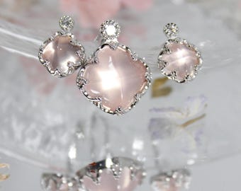 May include: A jewelry set featuring a pendant and two earrings. Each piece has a light pink, four-leaf clover-shaped stone set in a silver-colored metal. The earrings are smaller versions of the pendant design.