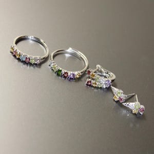 May include: A set of silver rings and earrings, each embellished with a variety of colourful gemstones. The rings display a line of small, bright stones, and the earrings feature a cluster of gems. The jewellery is set against a neutral backdrop.