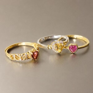 Peut inclure: Trois bagues ornées de pierres précieuses en forme de cœur. Une bague dorée présente un anneau en filigrane et une pierre rouge foncé. Une bague argentée possède une pierre jaune entourée de petites pierres claires. Une bague dorée a une pierre rose.