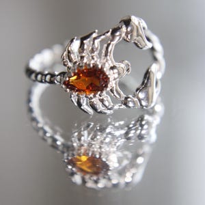 Può includere: Un anello d'argento con un design a scorpione. L'anello presenta un dettagliato scorpione con una gemma arancione al centro. L'anello è esposto su una superficie riflettente, mostrando il suo design e la sua maestria.