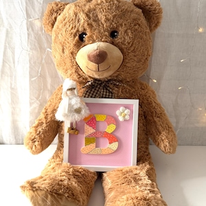 Puede incluir: Un gran oso de peluche marrón con una pajarita a cuadros. El oso sostiene una obra de arte enmarcada con un fondo rosa y la colorida letra "B". Una pequeña muñeca y una flor blanca también están unidas al marco.