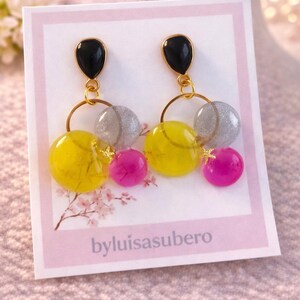 May include: A pair of dangle earrings with black teardrop-shaped tops, gold hoops, and clusters of yellow, silver, and pink beads. The earrings are displayed on a pink card with the text "byluisasubero".