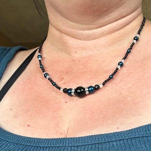 May include: A black beaded necklace with a large, round, black central bead. The necklace features smaller, dark blue and white beads interspersed along the black string. The necklace is worn around the neck.
