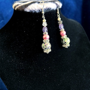 Pode incluir: Um par de brincos pendurados com ganchos prateados. Cada brinco apresenta uma combinação de contas rosa, roxas e transparentes, juntamente com um elemento floral decorativo. A parte inferior de cada brinco tem uma conta redonda estampada com um design de filigrana prateada.