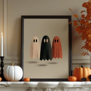 May include: A framed wall art print featuring three ghost figures in beige, black, and burnt orange colors. The ghosts have simple oval eyes and are positioned against a light tan background. The frame is black and rectangular in shape.