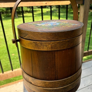 May include: A wooden bucket with a painted floral design on the lid and a wooden handle. The bucket is brown with a natural wood finish.