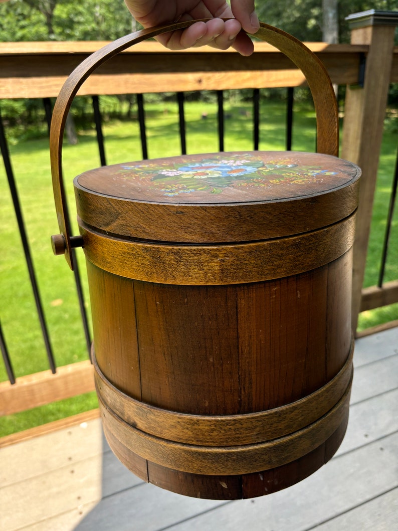 May include: A wooden bucket with a painted floral design on the lid and a wooden handle. The bucket is brown with a natural wood finish.