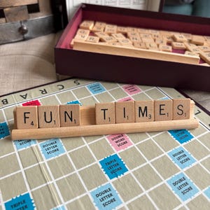 May include: A Scrabble game in progress, with the word "FUN TIMES" spelled out on the board. The wooden tiles are in a wooden rack, with the game board visible. The board has blue, pink, and green squares.