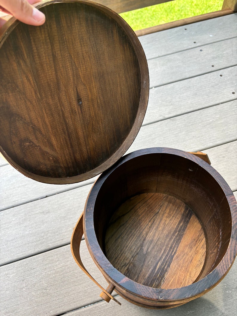 May include: A wooden bucket with a lid, the lid is open and the bucket is empty. The bucket has a brown wooden handle.