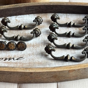 May include: A collection of antique-style brass hardware including six curved drawer pulls with ornate floral ends and three circular cabinet knobs featuring a central flower design, all arranged on a wooden tray with the word home printed on it.