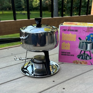 May include: A silver fondue pot with a black handle and lid, sitting on a metal stand with a heat source. The pot is on a wooden surface and the box for the fondue set is visible in the background. The box is pink with the words "2 Quart - all stainless steel fondue dish" and "heatproof handle adjustable flame control" printed on it. The box also has a picture of the fondue set on it.