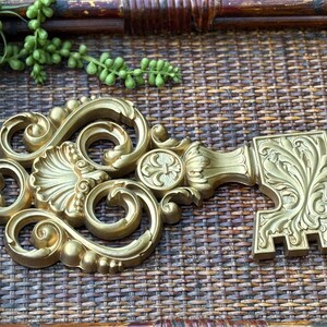 May include: Ornate gold-colored decorative key with intricate scrollwork and floral details. The key is lying on a woven brown surface, with green leafy sprigs in the upper left corner. A decorative accent piece.