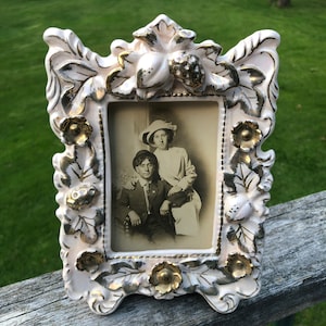 May include: A white porcelain picture frame with gold accents and floral details. The frame holds a black and white photograph of two people.
