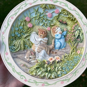 May include: A decorative ceramic plate with a relief scene of kittens and a mouse in a garden. The plate features a white border with a floral design and a detailed, colorful depiction of the animals among flowers and foliage.