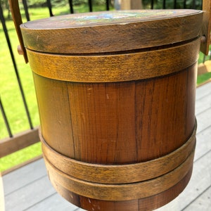 May include: A wooden bucket with a lid and a handle. The bucket is brown and has a natural wood finish. The lid has a painted design of flowers.