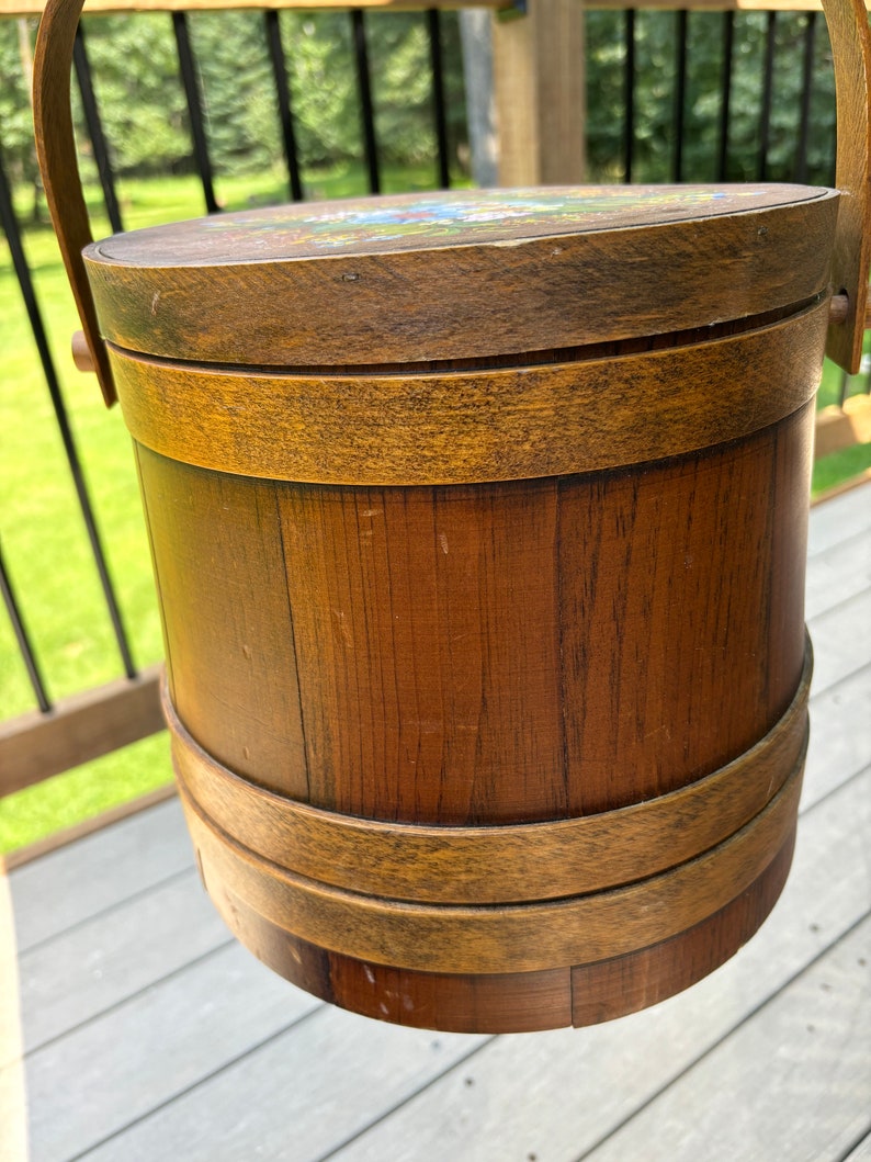 May include: A wooden bucket with a lid and a handle. The bucket is brown and has a natural wood finish. The lid has a painted design of flowers.