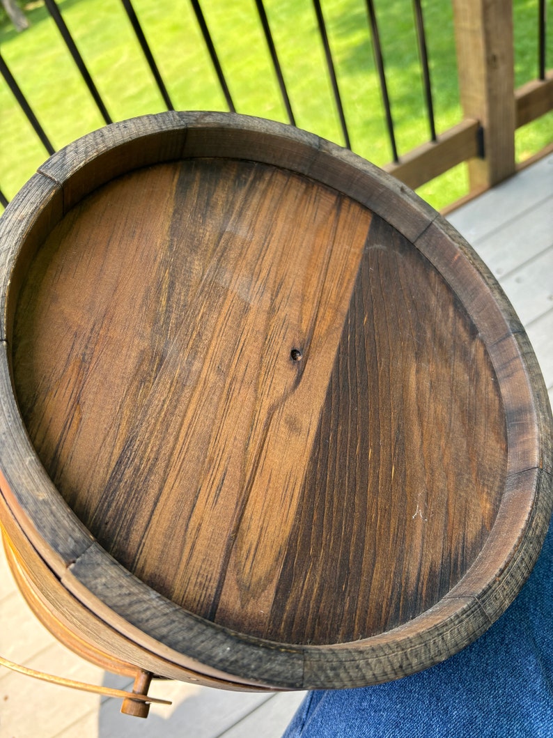 May include: A wooden bucket with a dark brown stain and a metal handle. The bucket is made of wood slats and has a round shape.