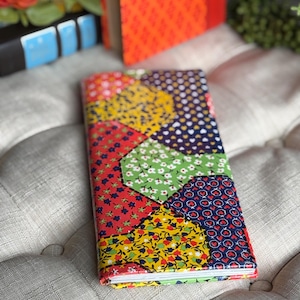 May include: A colorful journal with a patchwork design. The cover features various floral and heart patterns in red, yellow, green, and blue. The journal is resting on a tufted, light-colored surface, with other books in the background.