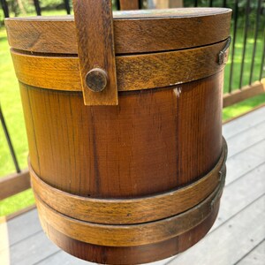 May include: A wooden bucket with a handle, featuring a brown stain and a natural wood grain. The bucket has two brown bands around the top and bottom.