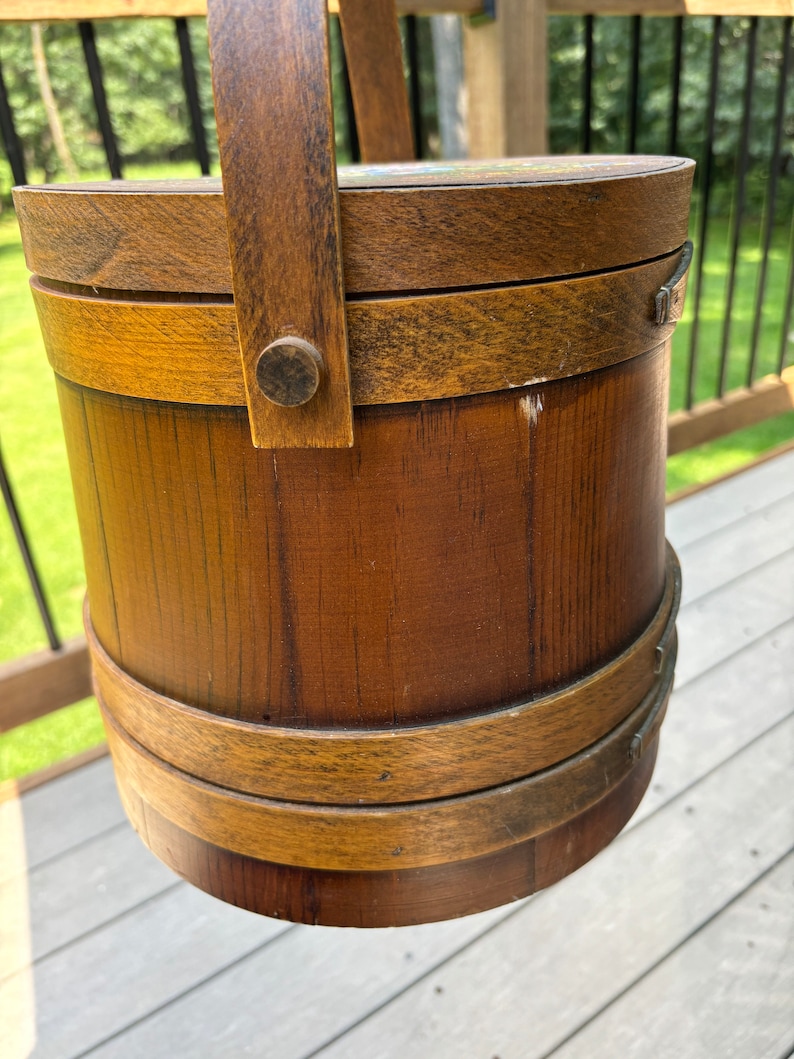 May include: A wooden bucket with a handle, featuring a brown stain and a natural wood grain. The bucket has two brown bands around the top and bottom.
