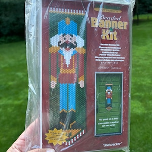 May include: A packaged "Nutcracker" beaded banner kit. The kit includes a full-size color pattern. The banner design features a nutcracker soldier in red, blue, and gold. The package dimensions are 6" x 23.75" (16x59cm).