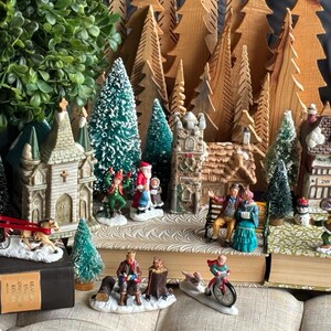 May include: A Christmas village scene with miniature houses, figurines, and trees. The village includes a church, houses, and people engaged in winter activities. The scene is set on books and decorated with artificial trees and greenery.