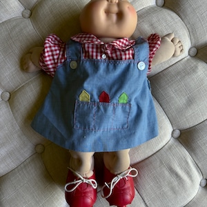 May include: A Cabbage Patch Kid doll wearing a red and white gingham shirt, blue overalls with a patch pocket, and red shoes.