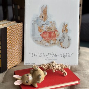 May include: A hardcover copy of "The Tale of Peter Rabbit" by Beatrix Potter, featuring an illustration of rabbits. A ceramic bunny figurine and a decorative cream-colored object rest on a red book.